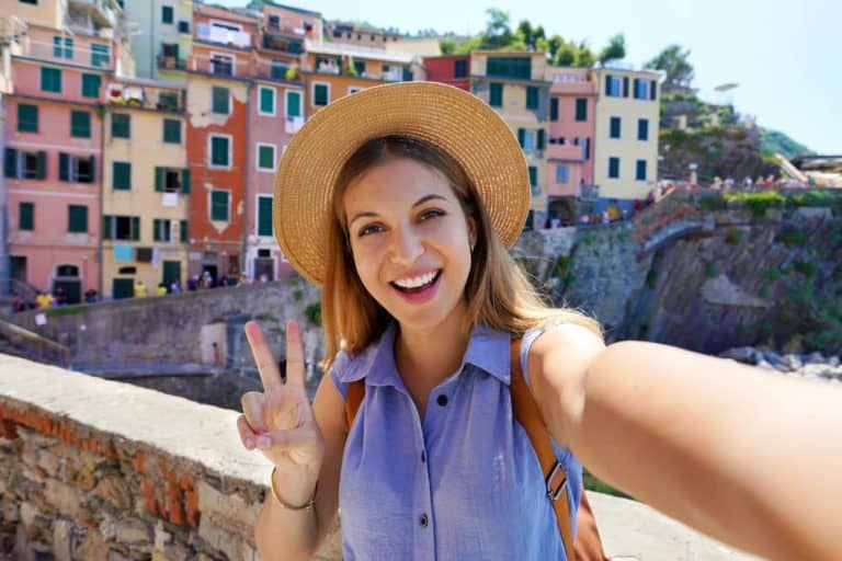 Happy traveler girl taking selfie photo doing victory sign on sunny day in Riomaggiore village, Cinque Terre, Italy